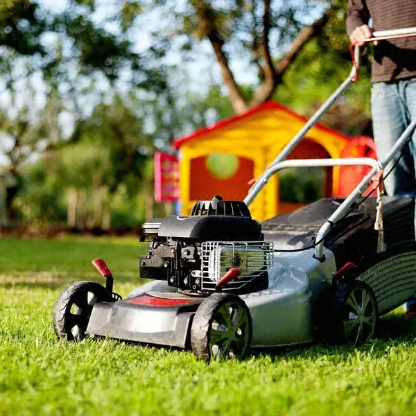 Push mower cutting thick green grass in a sunny backyard with a children's playhouse behind it