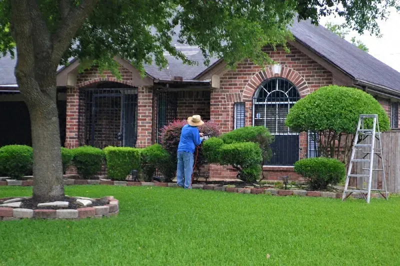 Worker trimming shrubs in front of a brick home with a green lawn